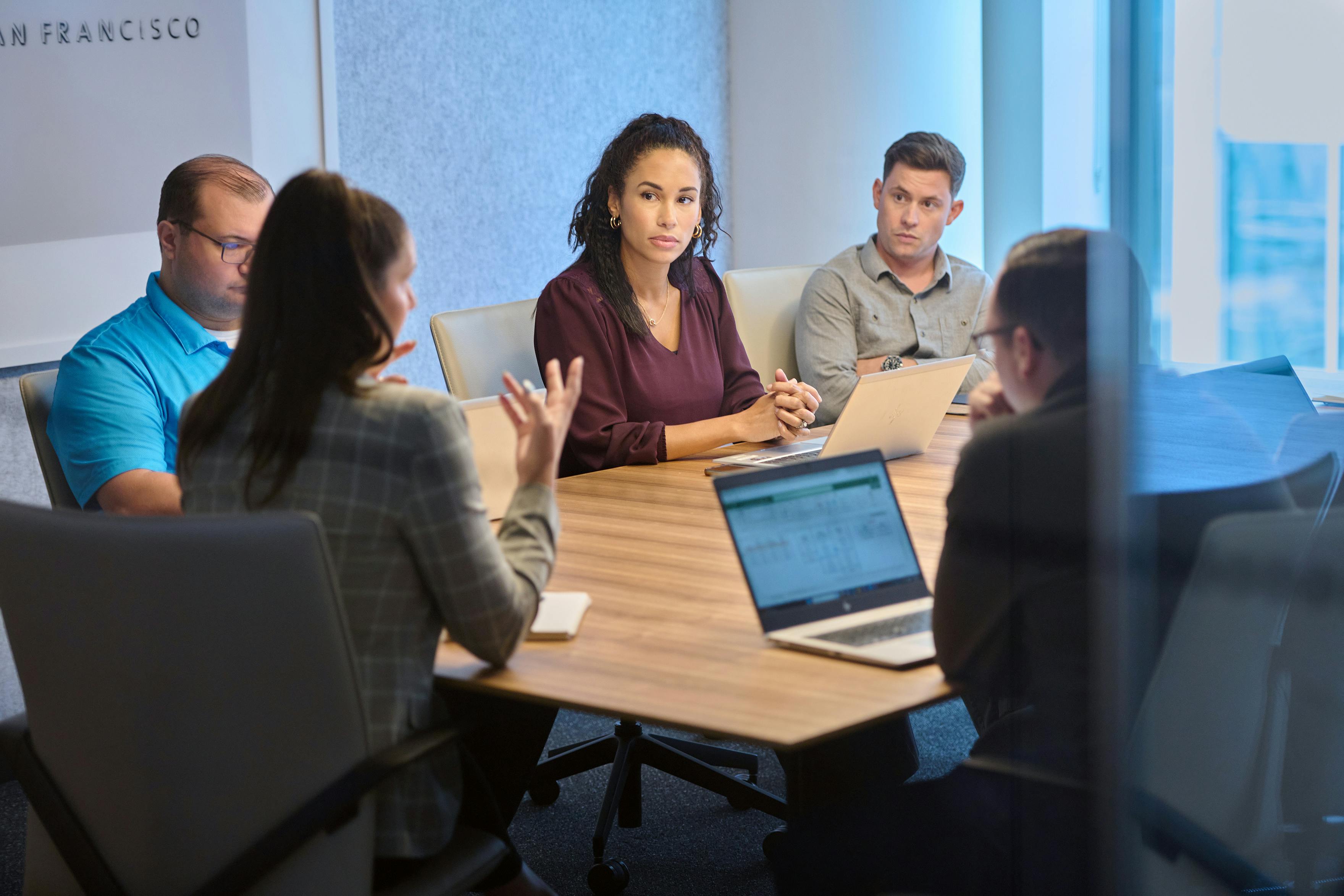 Employees are gathered in a small conference room, tightly packed around a central table. The employee at the head of the table gestures while presenting.